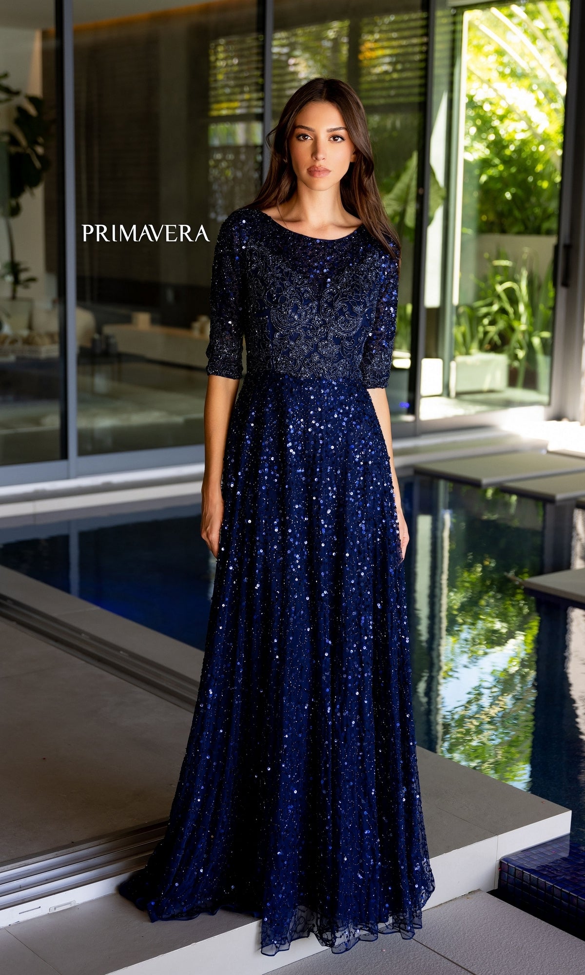 A woman wears the Long Prom Dress 13115 by Primavera, a navy blue sequined gown with a scoop neck and short sleeves. She stands indoors by a modern pool, greenery visible through large windows, the floor-length skirt creating an elegant silhouette.